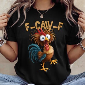 May include: Black t-shirt featuring a cartoon rooster with wide eyes and a long red tongue. The rooster has blue and brown feathers and is positioned below the text "F-CAW-F" in a yellow font. The shirt is a casual wear item.