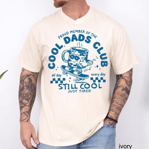 May include: Ivory-colored t-shirt with a blue graphic that reads "Cool Dads Club." The graphic features a coffee mug character on a skateboard, with the text "Still Cool Just Tired." The shirt has a v-neck collar.