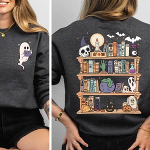 May include: A dark gray sweatshirt with a graphic of a bookshelf filled with books, a ghost, a cauldron, and other Halloween-themed items. The text "RIP" is visible on the bookshelf.