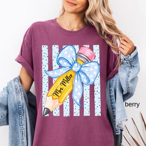 May include: A berry-colored t-shirt featuring a graphic of a yellow pencil with a pink eraser and a large light blue bow with white hearts. The pencil is printed with the text Mrs. Miller, set against a background of vertical light blue stripes.