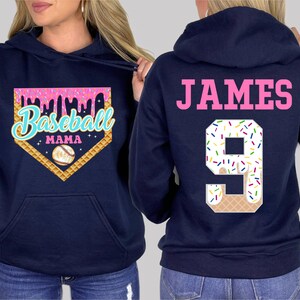 K&ouml;nnte beinhalten: Zwei marineblaue Kapuzenpullover. Ein Pullover hat ein pinkfarbenes und wei&szlig;es Baseball-Motiv mit dem Text "Baseball Mama". Der andere Pullover hat den Text "JAMES" und die Nummer "9" mit einem pinkfarbenen und wei&szlig;en Eiswaffel-Motiv.