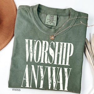May include: A moss green t-shirt with the words "WORSHIP ANYWAY" in large, distressed white letters. A gold-colored necklace with a small pendant rests on the shirt. The shirt is folded on a white surface.