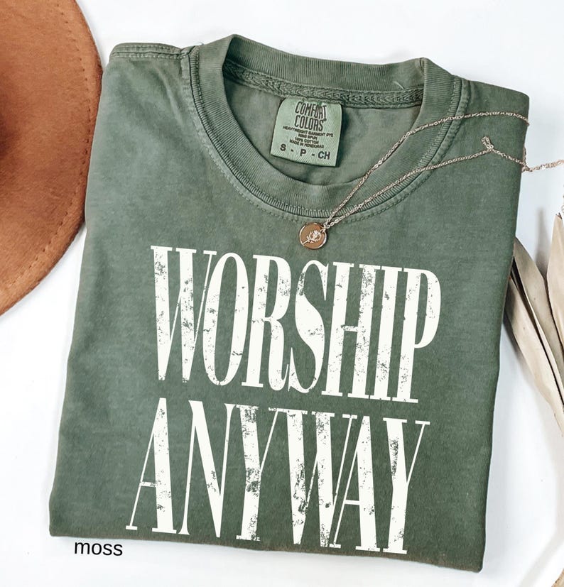 May include: A moss green t-shirt with the words "WORSHIP ANYWAY" in large, distressed white letters. A gold-colored necklace with a small pendant rests on the shirt. The shirt is folded on a white surface.