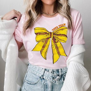 May include: Pink t-shirt with a yellow softball bow and the text "Your Team" on the front.