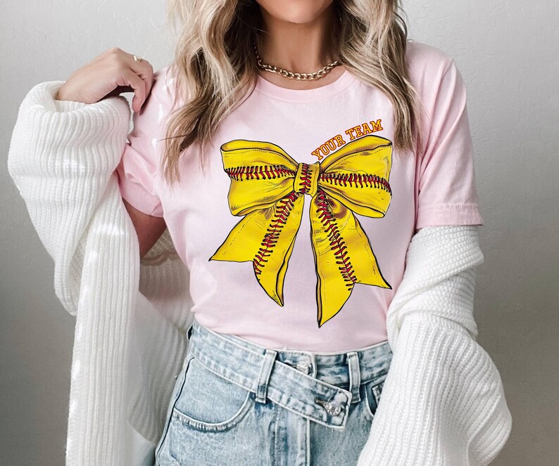 May include: Pink t-shirt with a yellow softball bow and the text "Your Team" on the front.