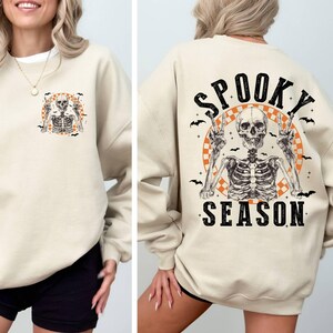 Puede incluir: Una sudadera beige con un gr&aacute;fico de esqueleto y el texto "Spooky Season" impreso en la espalda.