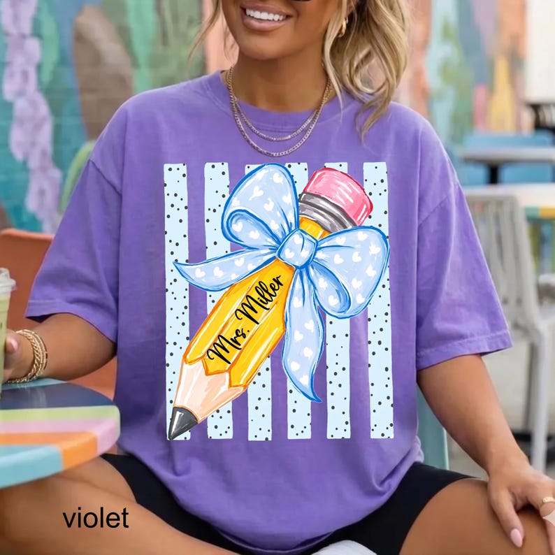 May include: A violet short-sleeved t-shirt featuring a graphic of a yellow pencil with a pink eraser and a large light blue bow with white hearts. The pencil has the text 'Mrs. Miller' written on it, set against a background of light blue vertical stripes with black dots.