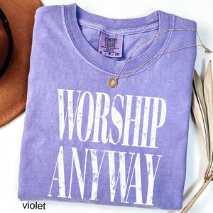 May include: A violet Comfort Colors t-shirt with the words "WORSHIP ANYWAY" in white block letters. A gold necklace with a pendant rests on the shirt. A brown hat and dried plant stems are visible in the background.