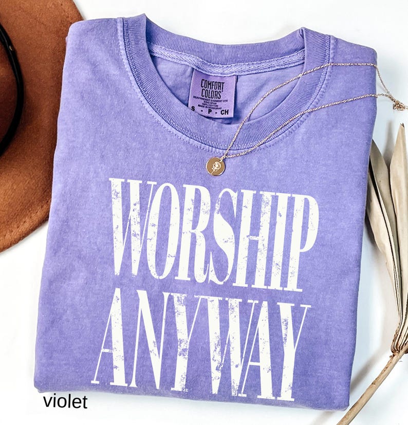 May include: A violet Comfort Colors t-shirt with the words "WORSHIP ANYWAY" in white block letters. A gold necklace with a pendant rests on the shirt. A brown hat and dried plant stems are visible in the background.