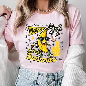 May include: Pink t-shirt with a cartoon banana wearing sunglasses, holding a baseball bat, and wearing sneakers. The shirt features the text "Bananas" and "Let's Go Bananas" in yellow, along with a baseball, a checkered bow, and a foam finger.