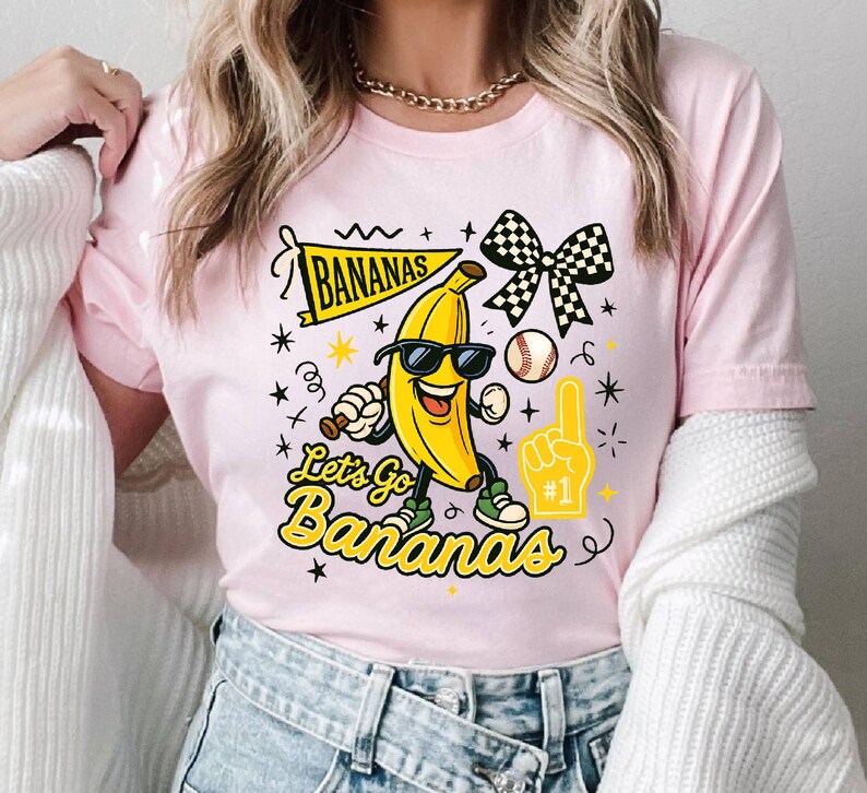 May include: Pink t-shirt with a cartoon banana wearing sunglasses, holding a baseball bat, and wearing sneakers. The shirt features the text "Bananas" and "Let's Go Bananas" in yellow, along with a baseball, a checkered bow, and a foam finger.