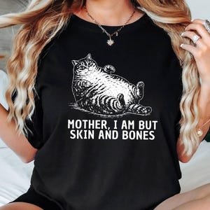 以下が含まれることがあります： 黒いTシャツ。白いイラストでリラックスした猫が描かれています。猫の下には「MOTHER, I AM BUT SKIN AND BONES」の文字。猫は仰向けに寝ており、足が上がっています。Tシャツは綿です。