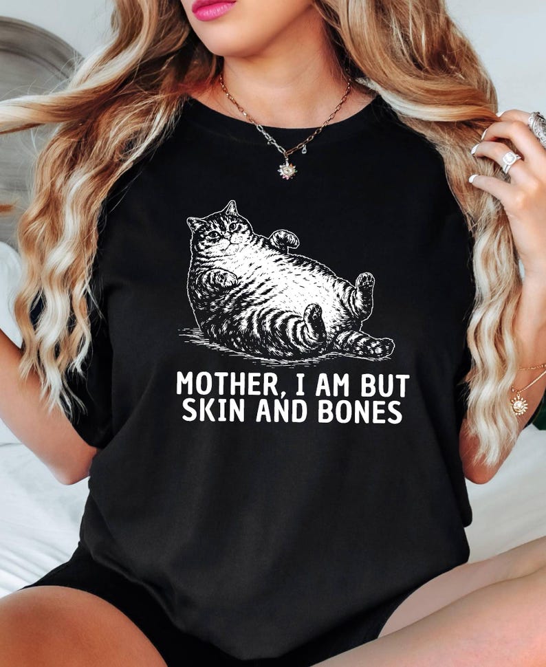 以下が含まれることがあります： 黒いTシャツ。白いイラストでリラックスした猫が描かれています。猫の下には「MOTHER, I AM BUT SKIN AND BONES」の文字。猫は仰向けに寝ており、足が上がっています。Tシャツは綿です。