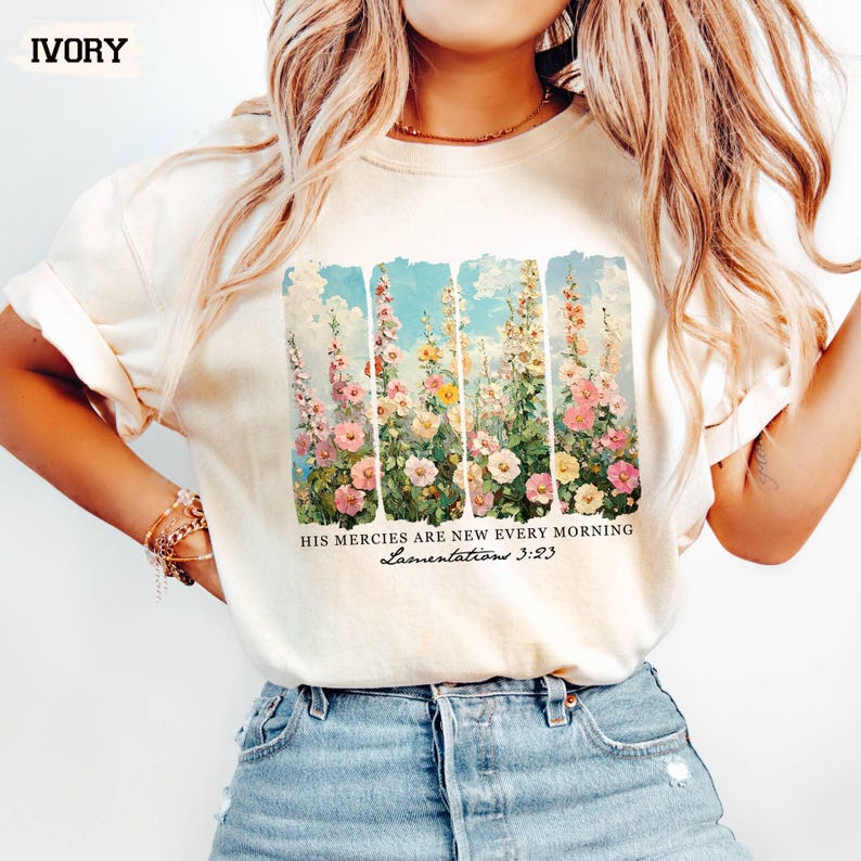 May include: Ivory-colored t-shirt featuring a floral design with the text "HIS MERCIES ARE NEW EVERY MORNING Lamentations 3:23." The design includes a variety of flowers in shades of pink, yellow, and white against a blue sky background.