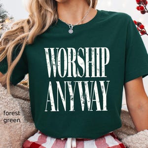 May include: Forest green t-shirt with the words "WORSHIP ANYWAY" in large, white, bold letters. The shirt is a solid color and has short sleeves. The model is wearing red and white plaid pajama pants.