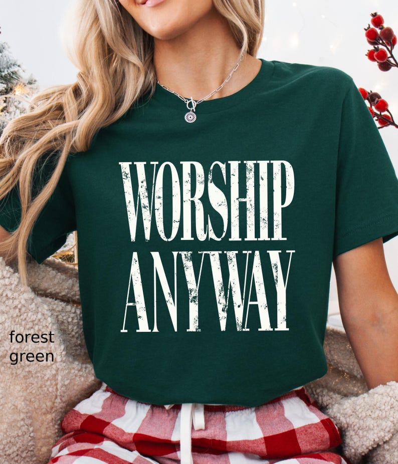 May include: Forest green t-shirt with the words "WORSHIP ANYWAY" in large, white, bold letters. The shirt is a solid color and has short sleeves. The model is wearing red and white plaid pajama pants.