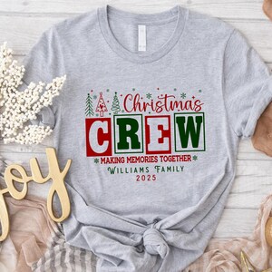 May include: A light grey t-shirt featuring a Christmas design. The graphic reads "Christmas CREW" in red and green, with the text "MAKING MEMORIES TOGETHER" and the family name "WILLIAMS FAMILY 2025".
