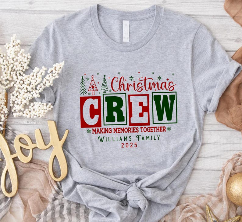 May include: A light grey t-shirt featuring a Christmas design. The graphic reads "Christmas CREW" in red and green, with the text "MAKING MEMORIES TOGETHER" and the family name "WILLIAMS FAMILY 2025".