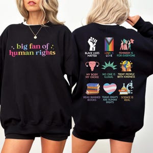 May include: Black sweatshirt with the text "big fan of human rights" in colorful letters on the front. The back features various graphics and phrases, including "Black Lives Matter," "Love is Love," and "Feminism is for Everyone."