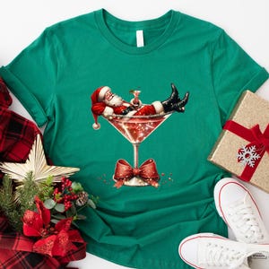 May include: A green t-shirt with a festive design featuring Santa Claus relaxing in a martini glass. The glass is filled with a red liquid and sparkles. Santa is wearing a red suit and a white beard.