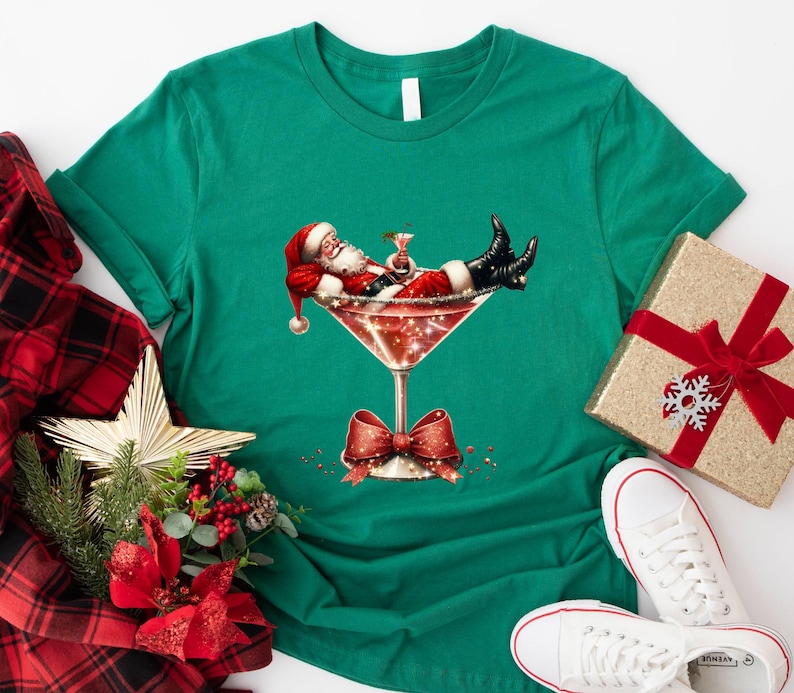May include: A green t-shirt with a festive design featuring Santa Claus relaxing in a martini glass. The glass is filled with a red liquid and sparkles. Santa is wearing a red suit and a white beard.