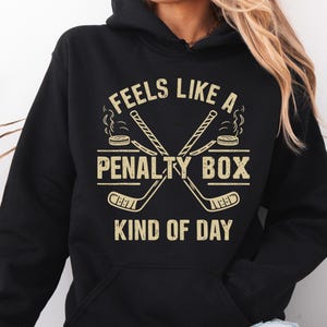 May include: Black hoodie with the text "FEELS LIKE A PENALTY BOX KIND OF DAY" in a distressed, beige font. The design includes crossed hockey sticks and a hockey puck with steam rising from it.