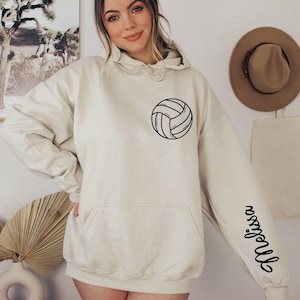 May include: A cream-colored hoodie with a black volleyball graphic on the front and the word "volleyball" written vertically on the right sleeve.