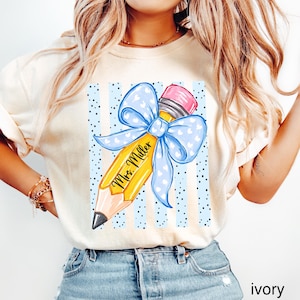 May include: An ivory t-shirt featuring a graphic of a yellow pencil with a large light blue bow patterned with white hearts. The pencil is inscribed with the text Mrs. Miller and is set against a background of vertical light blue stripes and black dots.
