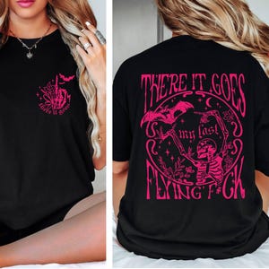 K&ouml;nnte beinhalten: Schwarzes T-Shirt mit einem rosafarbenen Grafikdruck eines Skeletts, das eine Fledermaus mit dem Text "There it goes my last flying f*ck" h&auml;lt.