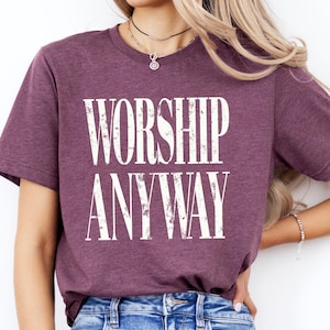 May include: A maroon t-shirt with the words "WORSHIP ANYWAY" printed in large, distressed white letters. The shirt is made of a soft material and has short sleeves. The model is wearing blue jeans.