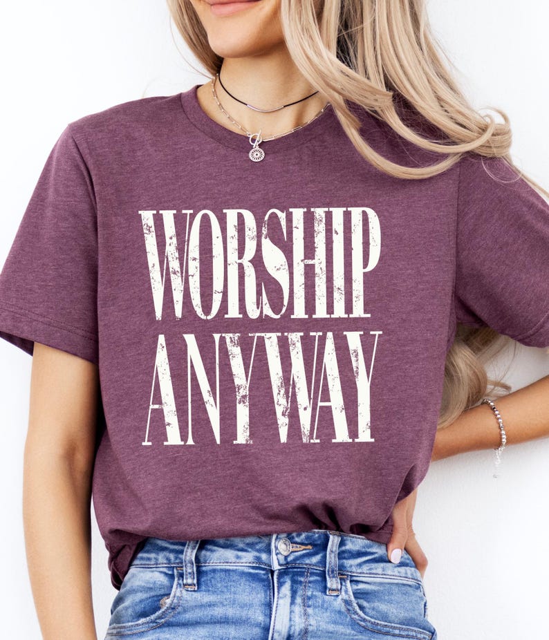 May include: A maroon t-shirt with the words "WORSHIP ANYWAY" printed in large, distressed white letters. The shirt is made of a soft material and has short sleeves. The model is wearing blue jeans.