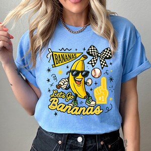 May include: Light blue t-shirt featuring a cartoon banana wearing sunglasses and holding a baseball bat. The shirt has the text "Bananas" and "Let's Go Bananas" in yellow, along with a baseball and a foam finger.