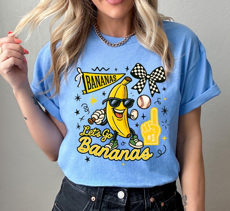 May include: Light blue t-shirt featuring a cartoon banana wearing sunglasses and holding a baseball bat. The shirt has the text "Bananas" and "Let's Go Bananas" in yellow, along with a baseball and a foam finger.