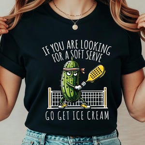 May include: Black t-shirt with a pickle cartoon playing pickleball. The pickle is wearing a headband and holding a yellow paddle. Text on the shirt reads "IF YOU ARE LOOKING FOR A SOFT SERVE GO GET ICE CREAM."