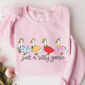 May include: A light pink sweatshirt featuring a whimsical design of four geese dressed in various outfits. The geese are adorned with hats, dresses, and a strawberry costume. The text "just a silly goose" is written below the geese in a playful font.