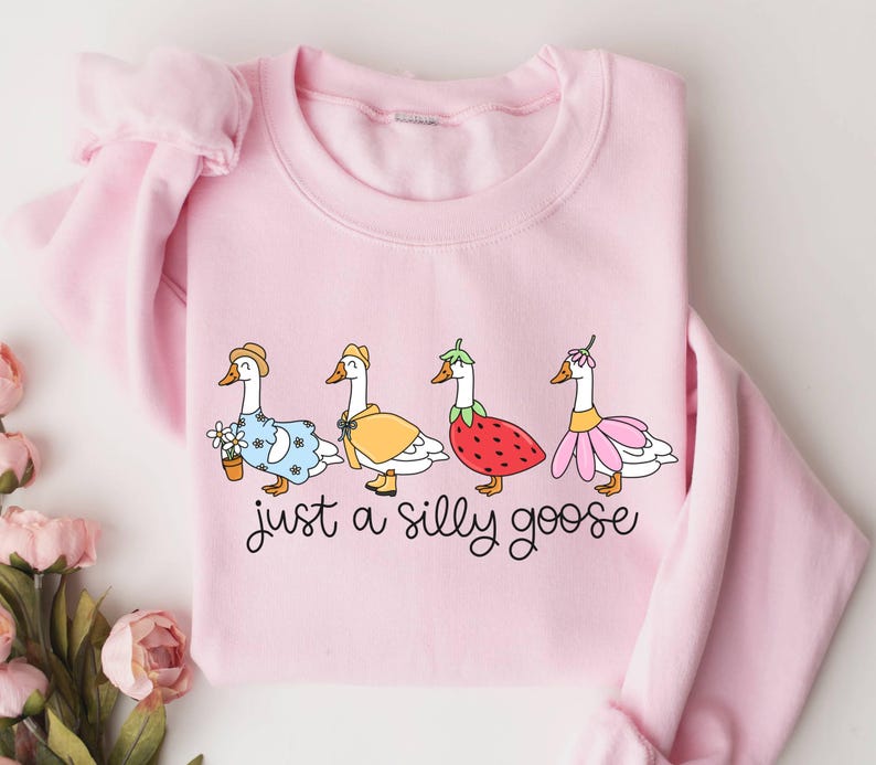 May include: A light pink sweatshirt featuring a whimsical design of four geese dressed in various outfits. The geese are adorned with hats, dresses, and a strawberry costume. The text "just a silly goose" is written below the geese in a playful font.