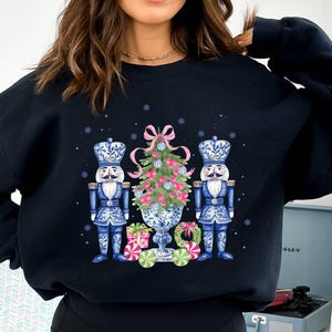 May include: A black sweatshirt with a Christmas-themed design featuring two blue and white nutcracker figurines flanking a decorated Christmas tree in a blue and white planter. The tree is adorned with pink and white ornaments and a pink bow.