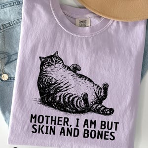 以下が含まれることがあります： 猫の黒いイラストが描かれた、蘭色のTシャツ。猫の下には「MOTHER, I AM BUT SKIN AND BONES」の文字がプリントされています。背景には、ベージュの帽子と青いジーンズがあります。