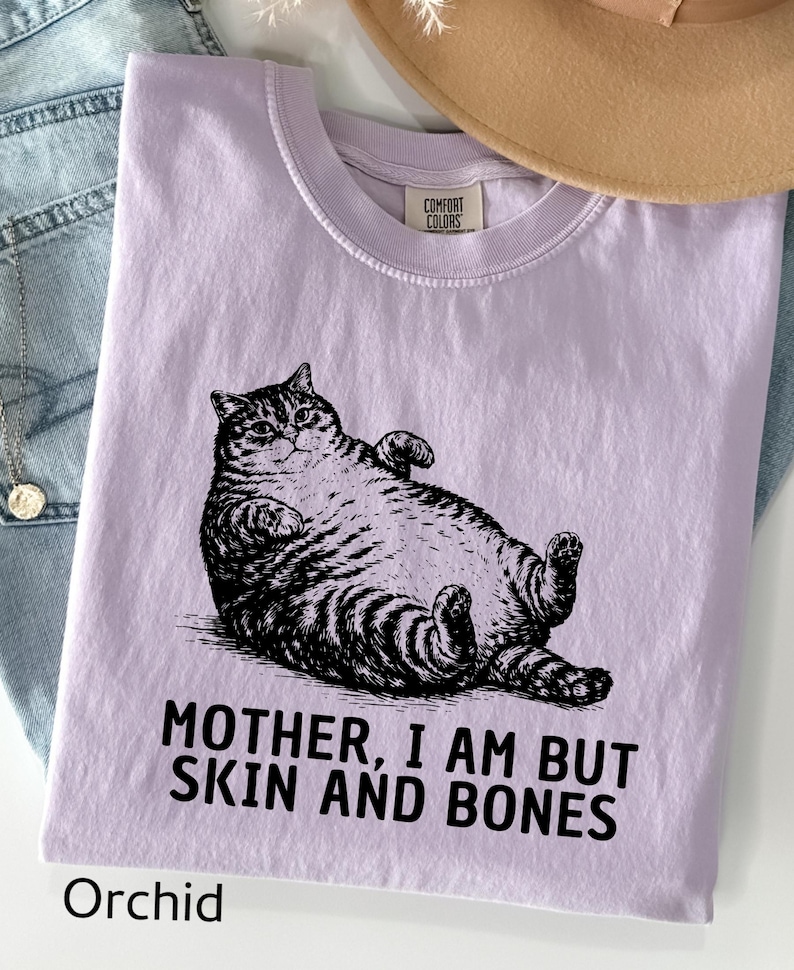以下が含まれることがあります： 猫の黒いイラストが描かれた、蘭色のTシャツ。猫の下には「MOTHER, I AM BUT SKIN AND BONES」の文字がプリントされています。背景には、ベージュの帽子と青いジーンズがあります。