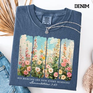 May include: Denim blue t-shirt with a floral design and the text "HIS MERCIES ARE NEW EVERY MORNING Lamentations 3:23". The shirt has a crew neck and a silver necklace. The shirt is made by Comfort Colors.