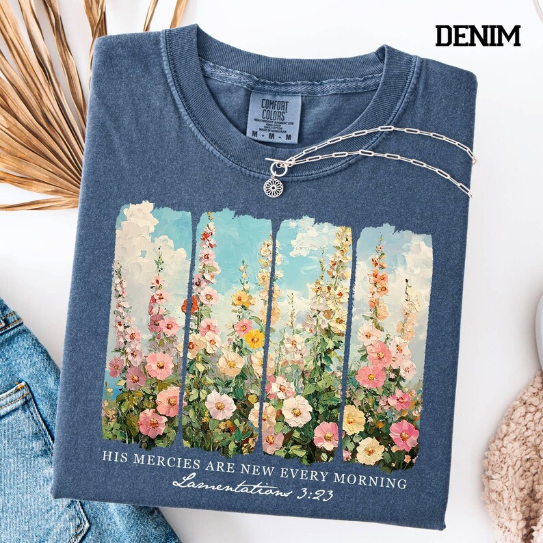 May include: Denim blue t-shirt with a floral design and the text "HIS MERCIES ARE NEW EVERY MORNING Lamentations 3:23". The shirt has a crew neck and a silver necklace. The shirt is made by Comfort Colors.