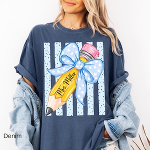 May include: A dark blue t-shirt featuring a graphic of a yellow pencil with a pink eraser and a large light blue bow with white hearts. The pencil is printed with the text Mrs. Miller, set against a background of vertical light blue stripes.