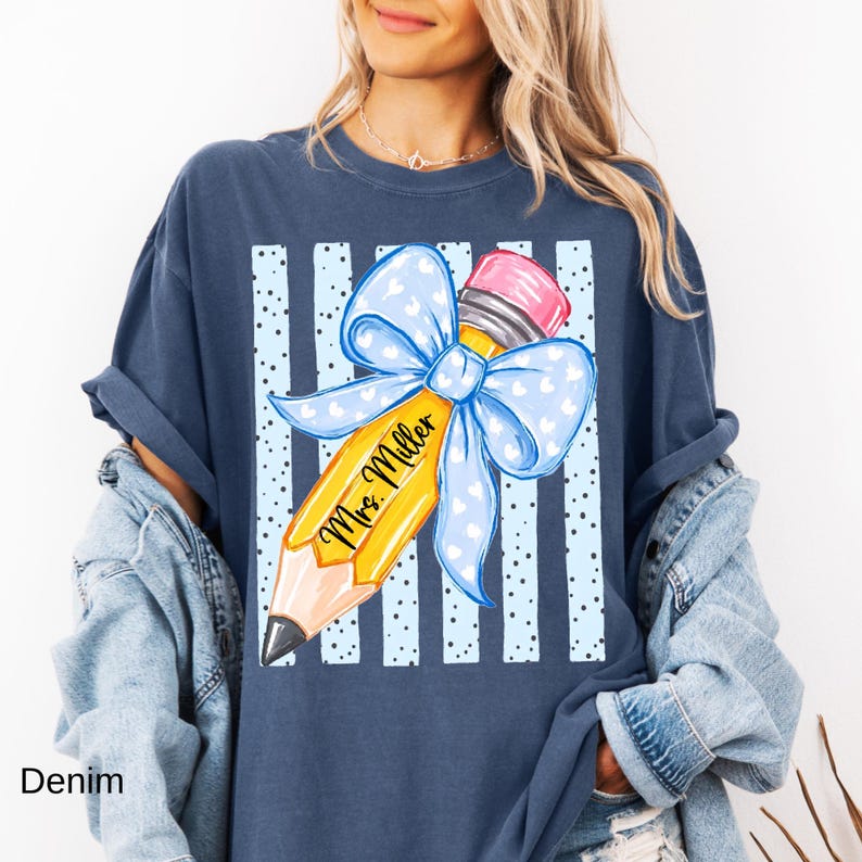 May include: A dark blue t-shirt featuring a graphic of a yellow pencil with a pink eraser and a large light blue bow with white hearts. The pencil is printed with the text Mrs. Miller, set against a background of vertical light blue stripes.