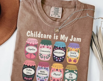 May include: A brown t-shirt with the text "Childcare is My Jam" and a graphic of nine jars of jam, each labeled with a different aspect of childcare, such as "Cognitive", "Communication", "Intellectual", "Social & Emotional", "Problem Solving", "Child's Development", "Physical", and "Life Skills".