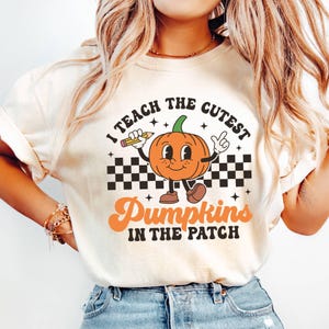May include: A white t-shirt with a graphic of a smiling pumpkin holding a pencil and the text "I teach the cutest pumpkins in the patch".