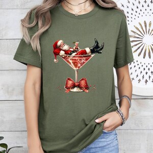 May include: A green t-shirt with a graphic of Santa Claus relaxing in a martini glass. The glass is decorated with a red bow and glitter.