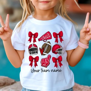 May include: White t-shirt with red and white cheerleading and football graphics. The shirt says "cheer" and "Your team name".