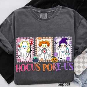 May include: Dark gray sweatshirt with a colorful graphic featuring three ghost characters dressed as medical professionals. The text "Hocus Poke-Us" is displayed in orange and purple. The design includes a syringe, a first aid kit, a cauldron, and a jack-o'-lantern.