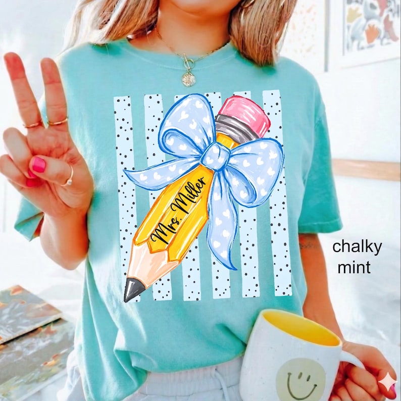 May include: A chalky mint t-shirt featuring a graphic of a yellow pencil with a pink eraser and a large light blue bow with white hearts. The pencil is printed with the text Mrs. Miller, set against a background of vertical white stripes.