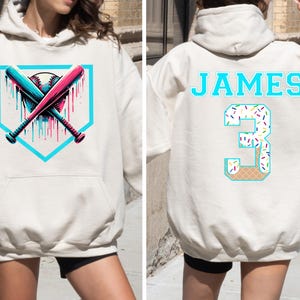 May include: Cream-colored hoodie with a baseball-themed graphic. The front features crossed bats and a baseball within a teal home plate shape. The back displays the name "JAMES" and the number "3" with a sprinkle and waffle cone design.